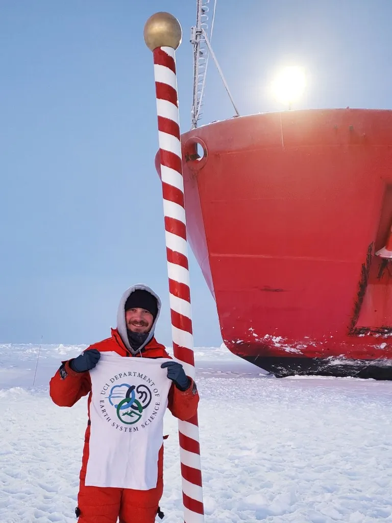 UCI graduate student Adam Fagan at the North Pole holding a UCI Earth System Science shirt beside a research ship
