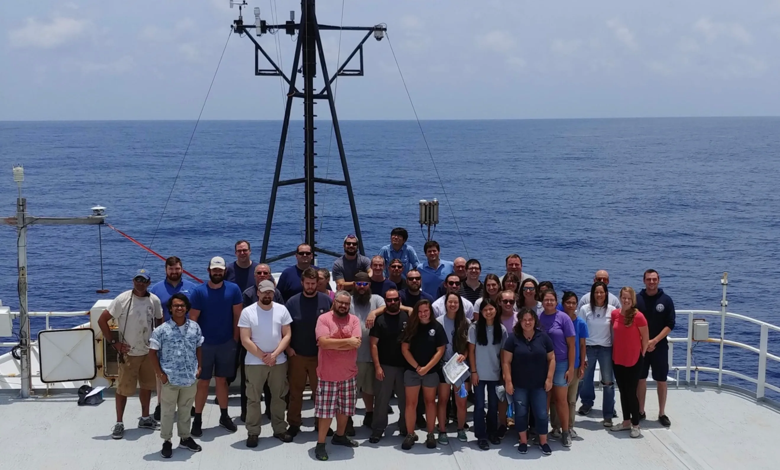 Group photo of about forty scientists and crew standing on the ship's deck in front of a mast, with calm blue ocean behind them
