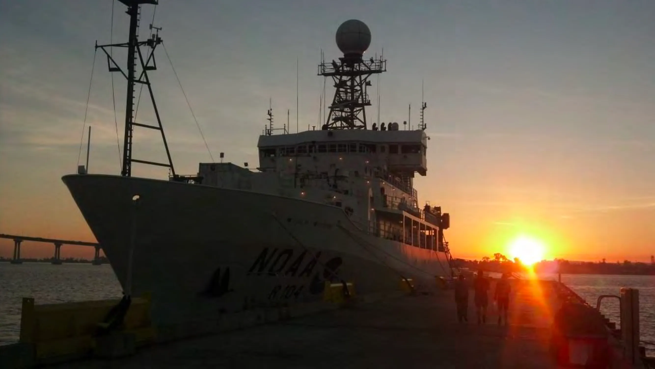The NOAA ship Ronald H. Brown silhouetted at a pier at sunset, with a bridge in the background